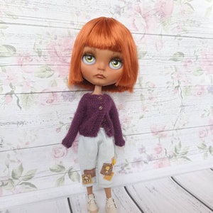 Puede incluir: Una mu&ntilde;eca con el pelo naranja, ojos verdes y un c&aacute;rdigan morado. La mu&ntilde;eca lleva pantalones blancos con parches marrones y zapatos blancos.
