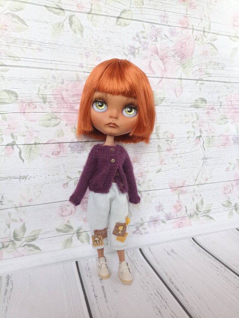 Puede incluir: Una mu&ntilde;eca con el pelo naranja, ojos verdes y un c&aacute;rdigan morado. La mu&ntilde;eca lleva pantalones blancos con parches marrones y zapatos blancos.