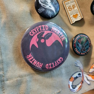 May include: A round button with the text "CRYPTID HUNTER" in pink, encircling a pink moth-like creature on a dark gray background. Other pins with various designs are visible in the background.