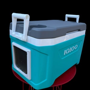 K&ouml;nnte beinhalten: Eine t&uuml;rkisfarbene und wei&szlig;e Igloo-K&uuml;hlbox mit einem grauen Deckel und einer integrierten Haustierklappe. Die K&uuml;hlbox hat zwei wei&szlig;e Griffe und einen Getr&auml;nkehalter auf dem Deckel.