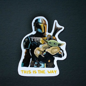 Op de afbeelding: Een witte sticker met een cartoon afbeelding van een Mandalorian-personage dat een klein groen buitenaards wezen vasthoudt. De tekst "This is the Way" is in geel gedrukt aan de onderkant van de sticker.