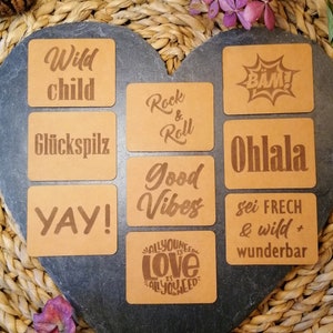 Könnte beinhalten: Neun braune Lederuntersetzer mit verschiedenen Sprüchen und Designs, darunter "Wild child", "Glückspilz", "Rock & Roll", "BAM", "Ohlala", "Good Vibes", "YAY", "All you need is love" und "Sei frech & wild + wunderbar".