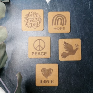 Könnte beinhalten: Fünf braune quadratische Untersetzer mit verschiedenen Designs und Wörtern. Die Untersetzer zeigen die Wörter "Love Peace Joy", "Hope", "Peace", "Love" und eine Taube mit einem Olivenzweig.