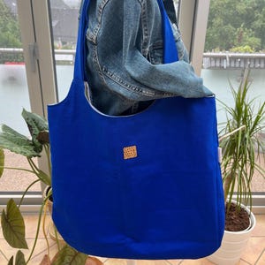 Könnte beinhalten: Eine große, königsblaue Tragetasche mit langen Trägern. Die Tasche hat einen abgerundeten Boden und einen kleinen, quadratischen, gemusterten Aufnäher auf der Vorderseite. Die Tasche wird auf einer weißen Schaufensterpuppe präsentiert, mit einer Jeansjacke dahinter. Der Hintergrund umfasst Pflanzen und ein Fenster.