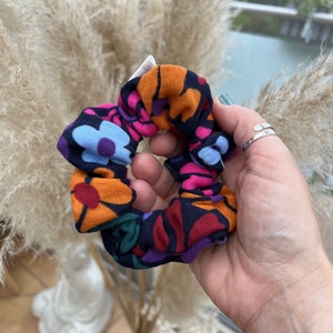 Könnte beinhalten: Ein farbenfrohes Haargummi mit einem Blumenmuster in Orange, Pink, Blau und Lila auf dunklem Hintergrund. Das Haargummi wird in einer Hand gehalten, was sein lebendiges Design zeigt. Ein Haaraccessoire.