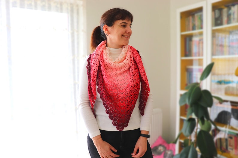 May include: A woman wearing a white shirt and black pants is standing in front of a window. She is wearing a crocheted shawl with a gradient of pink, red, and burgundy colors.
