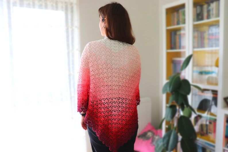 May include: A woman wearing a crocheted shawl with a gradient of red, pink, and white colors. The shawl is made with a delicate openwork pattern.