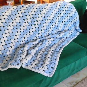 Könnte beinhalten: Handgefertigte Häkeldecke im Granny-Square-Muster mit abwechselnden weißen und hellblauen Quadraten. Die Decke liegt auf einem grünen Sofa. Eine schwarze Katze sitzt auf dem Sofa.