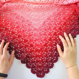 May include: A crocheted shawl with a gradient of pink, red, and burgundy colors. The shawl is made with a delicate lace pattern and has a scalloped edge.