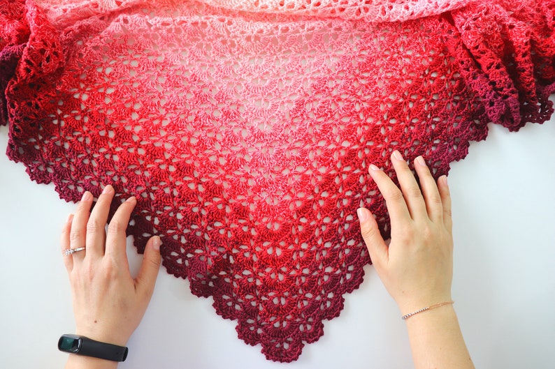 May include: A crocheted shawl with a gradient of pink, red, and burgundy colors. The shawl is made with a delicate lace pattern and has a scalloped edge.