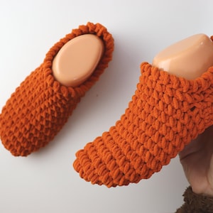 Puede incluir: Un par de zapatillas de punto naranja. Las zapatillas están hechas de un hilo grueso y texturizado y tienen una punta redondeada. Una zapatilla está sobre una forma de pie, y la otra está plana. Las zapatillas son de un estilo cálido y acogedor.