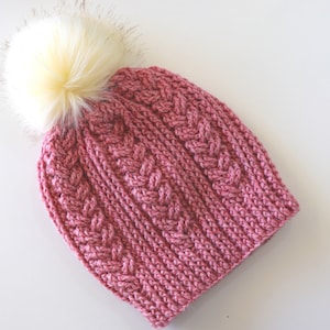 以下が含まれることがあります： 白いフェイクファーのポンポンが付いたピンクのニット帽。帽子はケーブルニットの模様です。