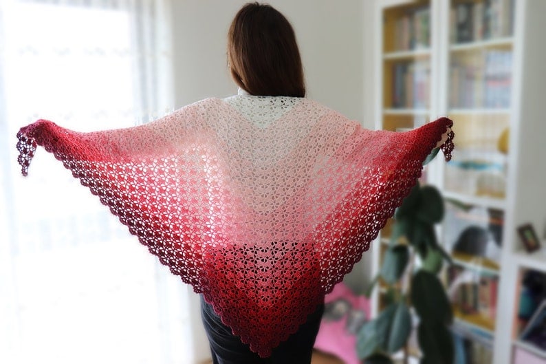 May include: A crocheted shawl with a gradient of pink, red, and white colors. The shawl is made with a delicate lace pattern and has a scalloped edge.
