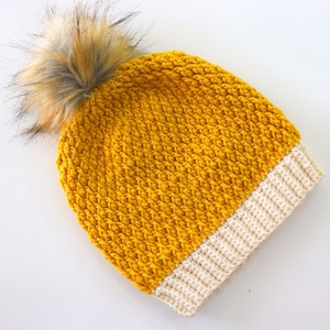 May include: A yellow crocheted beanie with a white brim and a brown faux fur pom pom.