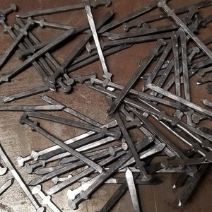 May include: A pile of silver metal swords with a rounded tip and a cross guard. The swords are arranged in a random pattern on a brown surface.
