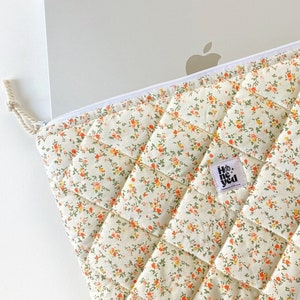 May include: A quilted laptop sleeve with a floral pattern in shades of orange, yellow, and green. The sleeve has a zipper closure and a small label that says "Hello, you".