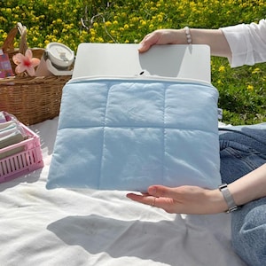 May include: A light blue quilted laptop sleeve with a white interior. The sleeve is held open, revealing a laptop. A picnic basket, pink container, and other items are nearby on a white blanket, set against a backdrop of yellow flowers.