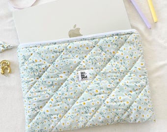 May include: A light blue quilted laptop sleeve with a white zipper and a daisy floral pattern. The sleeve is open, revealing a silver laptop. A small tag with text is visible on the sleeve.