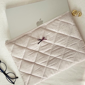 May include: A beige laptop sleeve with a quilted pattern and small black polka dots. A small burgundy bow is sewn on the front of the sleeve.