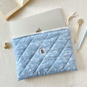 May include: A light blue quilted laptop sleeve with a white floral design. The sleeve has a white zip and a small tag with the text "He. You". A laptop is partially visible inside. A gold pen and a light blue pen are next to the sleeve.