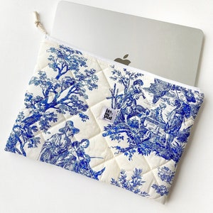 May include: A white quilted laptop sleeve with a blue and white toile pattern featuring a pastoral scene. The sleeve has a small white label with the text "He's the" on it.