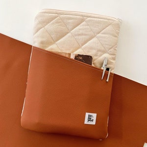 以下が含まれることがあります： ベージュのキルティングフラップが付いた茶色の革製のジッパーポーチ。ポーチには、前面に「He & She」と書かれた白いラベルが付いた小さなポケットがあります。