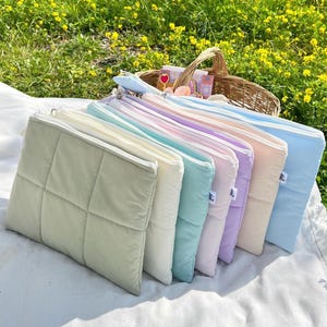 Pode incluir: Uma coleção de bolsas acolchoadas em tons pastel, dispostas sobre uma manta branca. As bolsas são em tons de verde, branco, azul, roxo e pêssego. Uma cesta de vime cheia de itens está no fundo, com um campo de flores amarelas.