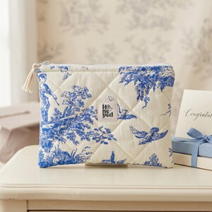 Könnte beinhalten: Eine gesteppte Kosmetiktasche mit blau-weißem Toile-de-Jouy-Muster mit Figuren, Bäumen und Vögeln. Die Tasche hat einen weißen Reißverschluss und ein kleines Etikett mit dem Text "He. Yed". Eine Karte mit dem Wort "Glückwunsch" ist ebenfalls sichtbar.