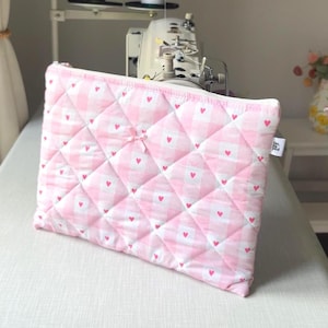 May include: A pink and white quilted pouch with a gingham pattern and small red hearts. The pouch has a zip closure and a small bow. The background includes a sewing machine and spools of thread.