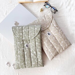 Op de afbeelding: Twee gewatteerde laptophouders met bloempatronen. Eén is saliegroen, de andere beige. Beide hebben een klepsluiting en een klein label. Een laptop, bril en accessoires zijn ook zichtbaar.