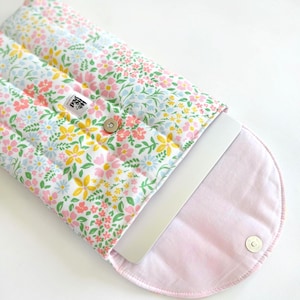 May include: Laptop sleeve with a floral pattern in white, pink, yellow, and blue. The quilted sleeve has a pink interior. A silver laptop is partially visible. The sleeve has a magnetic closure. The label reads "Hed".