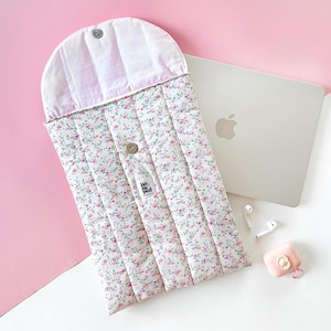 May include: A quilted laptop sleeve with a floral pattern. The sleeve has a white base with pink and green floral details. It features a rounded flap secured with a button. A silver laptop and accessories are next to the sleeve.