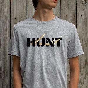 May include: A heather gray t-shirt with the word "HUNT" in black block letters. The letter "U" is replaced with a gold-colored antler design, and a gold antler design is also on the right side of the letter "T".