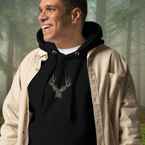 K&ouml;nnte beinhalten: Ein schwarzer Kapuzenpullover mit einem Tarnmuster-Hirsch-Silhouetten-Motiv. Das Model tr&auml;gt den Pullover &uuml;ber einem beigefarbenen Hemd mit Kn&ouml;pfen.