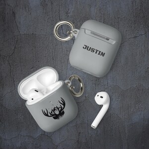 May include: Two gray Airpod cases with a keychain. One case has the name "JUSTIN" printed on it. The other case has a black silhouette of a deer head with antlers and mountains.
