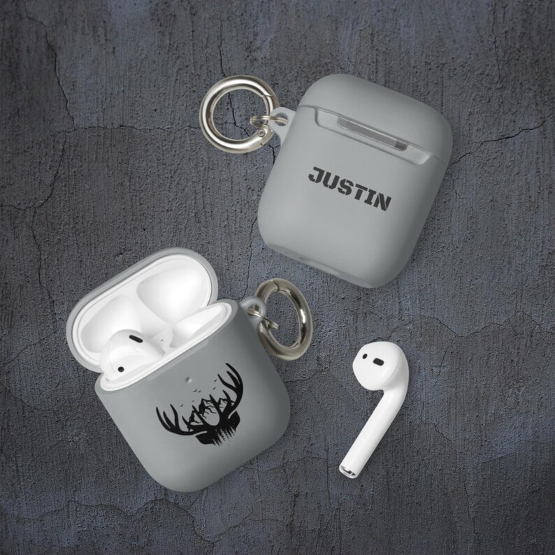 May include: Two gray Airpod cases with a keychain. One case has the name "JUSTIN" printed on it. The other case has a black silhouette of a deer head with antlers and mountains.