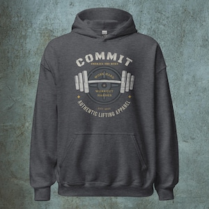 May include: A dark grey hooded sweatshirt with a white graphic design featuring a barbell and the text "COMMIT EMBRACE THE BURN WORK HARD WORKOUT HARDER AUTHENTIC LIFTING APPAREL EST. 2024".