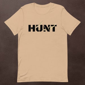 May include: A tan t-shirt with the word "HUNT" printed in black block letters. The letter "U" is partially obscured by a gold-colored design. The shirt is made of a soft material and has short sleeves.