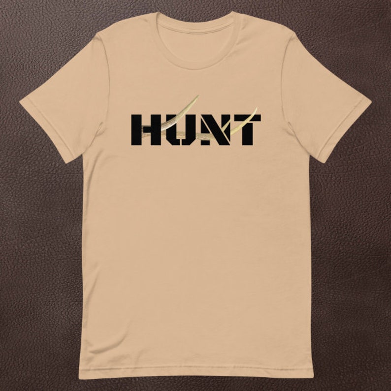 May include: A tan t-shirt with the word "HUNT" printed in black block letters. The letter "U" is partially obscured by a gold-colored design. The shirt is made of a soft material and has short sleeves.