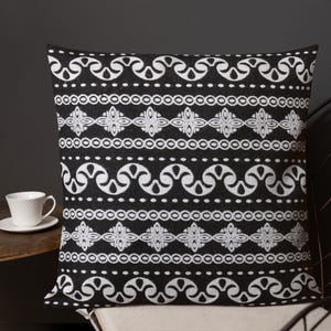 May include: Black and white patterned throw pillow. The pillow features a repeating design of white scroll and geometric shapes on a black background. The pillow is square and appears to be made of a soft fabric.