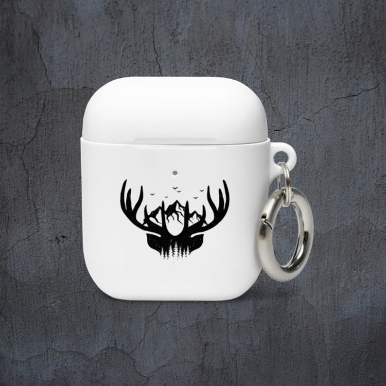 May include: White Airpods case with a black silhouette of a deer head with antlers and mountains in the background. The case has a silver metal ring attached to it.