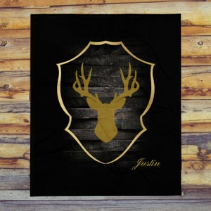May include: Black and gold throw blanket with a deer head silhouette inside a shield shape. The shield has a wood grain texture. The text "Justin" is at the bottom of the blanket.