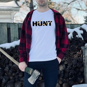 May include: White t-shirt with the word "HUNT" in black block letters, bisected by two gold-colored lines. The person is wearing a red and black plaid flannel shirt and black pants. An axe is held in the person's hand.