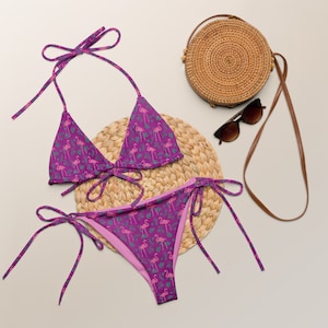 May include: A purple bikini with a pink flamingo print. The bikini top is a triangle style with adjustable straps. The bikini bottom is a cheeky style with ties on the sides. The bikini is on a woven straw mat.