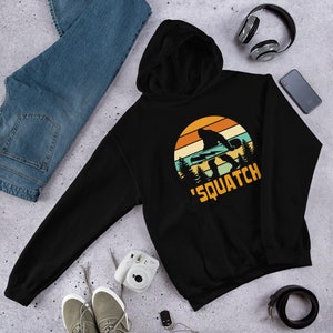 May include: Black hooded sweatshirt with a retro sunset graphic featuring a Bigfoot holding a snowboard. The text 'Squatch' is printed below the graphic.