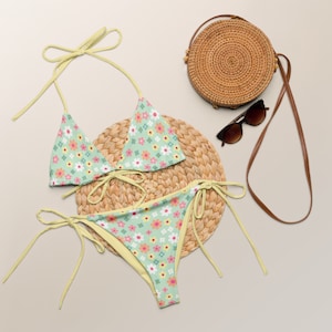 May include: A green and yellow floral print bikini with a matching straw bag and sunglasses. The bikini top is a triangle style with adjustable straps and the bottom is a high-waisted style with ties on the sides.