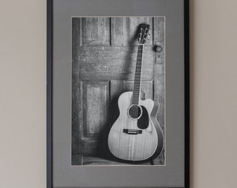 May include: A black and white photograph of an acoustic guitar leaning against a wooden door.