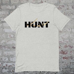 May include: Light gray t-shirt with the word "HUNT" in black block letters. The "U" and "N" are filled with a gold-colored pattern. Gold-colored antler-like shapes are above the "H" and "T".
