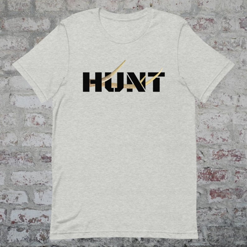 May include: Light gray t-shirt with the word "HUNT" in black block letters. The "U" and "N" are filled with a gold-colored pattern. Gold-colored antler-like shapes are above the "H" and "T".
