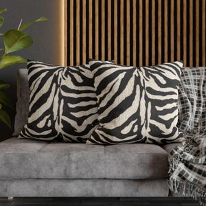 May include: Two square throw pillows with a black and cream zebra stripe pattern. The pillows are on a gray sofa with a plaid blanket. The background features a wooden slat wall and a green plant.